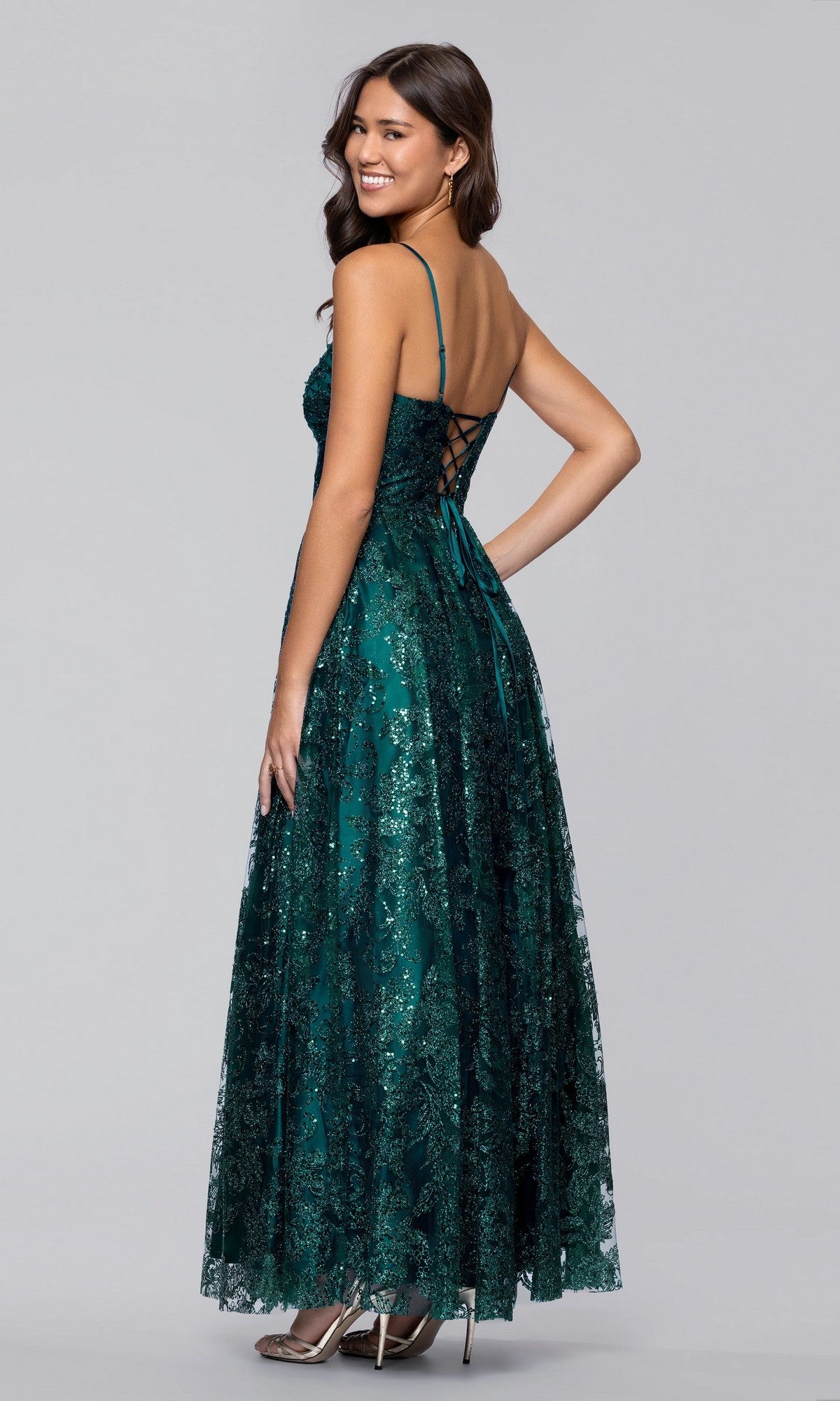 A woman in the Blondie Nites 5409BN long prom dress, featuring a sleeveless dark green glitter design with a lace-up corset bodice and sparkling embellishments, stands smiling over her shoulder.